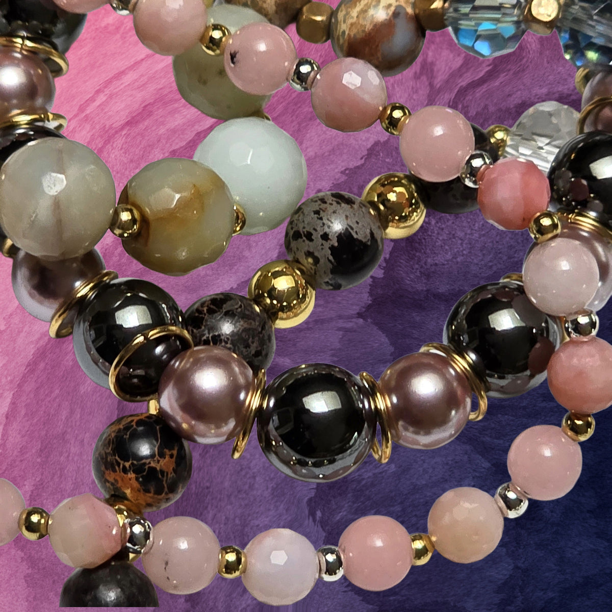 Close-up of a beaded strand jewelry piece featuring round beads in pink, black, white, and brown agate-like stone, accented with gold and silver spacer beads, set against a purple fabric background.